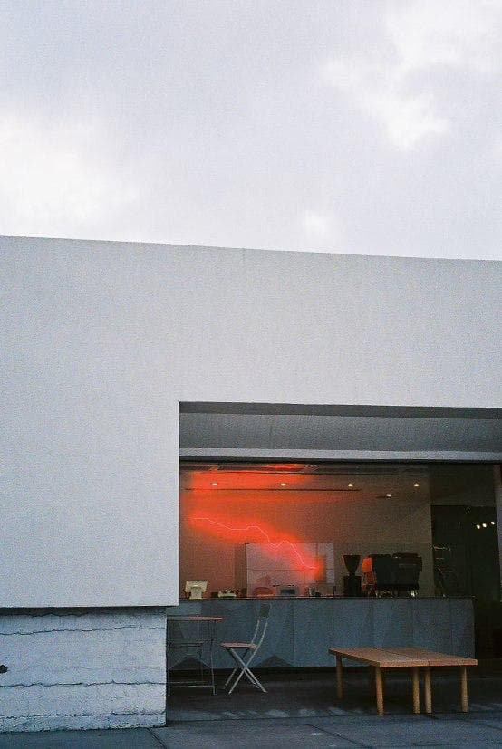 Minimal white building facade with an open doorway, warm interior light and a red neon line on the wall